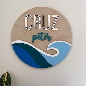 May include: A round wooden sign with the name "CRUZ peter" written in white and teal lettering. The sign features a blue and teal wave design.