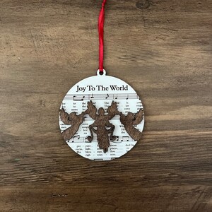 May include: A white round Christmas ornament with a brown wooden cutout of a moose and the words "Joy To The World" printed on it. The ornament is hanging from a red ribbon.