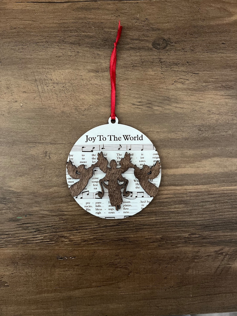 May include: A white round Christmas ornament with a brown wooden cutout of a moose and the words "Joy To The World" printed on it. The ornament is hanging from a red ribbon.