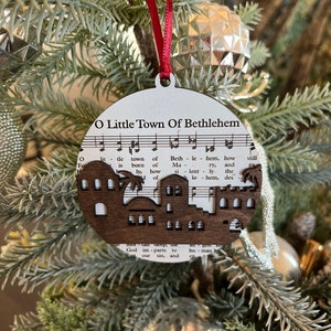 May include: A wooden Christmas ornament shaped like a town with the words "O Little Town of Bethlehem" printed on it. The ornament is decorated with a musical score and a silhouette of a town.