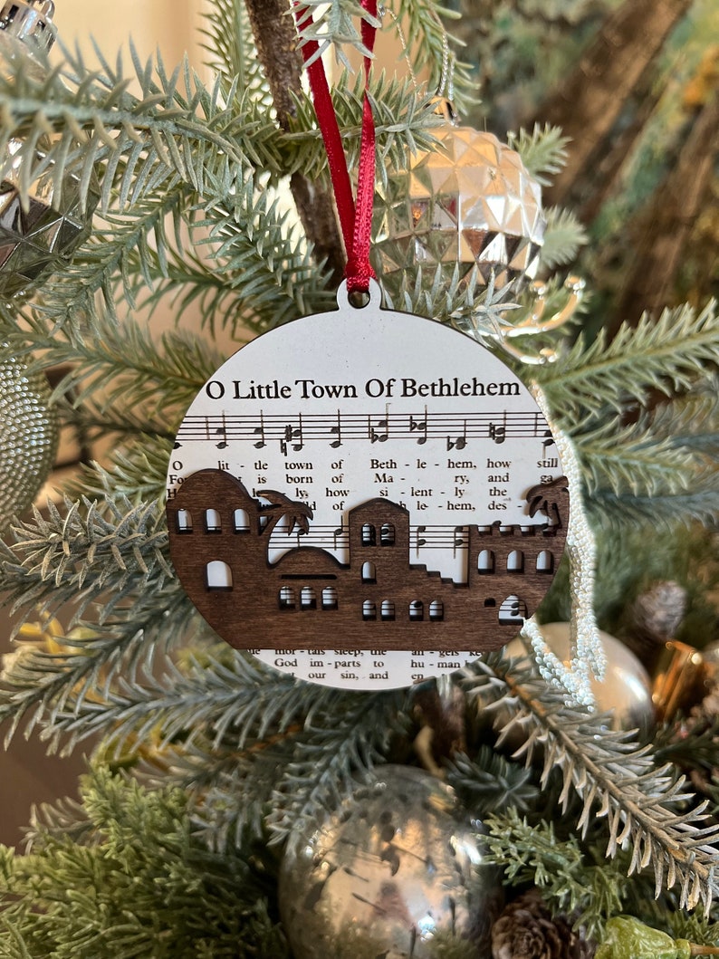 May include: A wooden Christmas ornament shaped like a town with the words "O Little Town of Bethlehem" printed on it. The ornament is decorated with a musical score and a silhouette of a town.