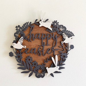 Pode incluir: Um sinal redondo de madeira com as palavras "happy easter" em letras de escrita preta. O sinal é decorado com uma coroa de flores, folhas, pássaros e coelhos recortados em papel preto.
