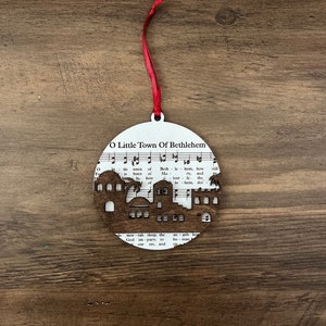 May include: A white wooden ornament shaped like a circle with a cutout of a town silhouette. The ornament features the words "O Little Town of Bethlehem" and music notes.