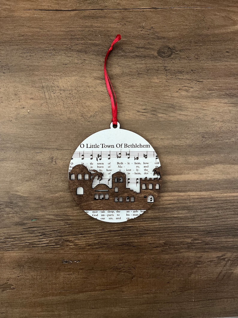 May include: A white wooden ornament shaped like a circle with a cutout of a town silhouette. The ornament features the words "O Little Town of Bethlehem" and music notes.