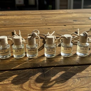 May include: A collection of small, clear glass bottles with wooden caps and attached twine cords. Each bottle has a label that reads "Main Street 808 HI". The bottles are arranged on a wooden surface.