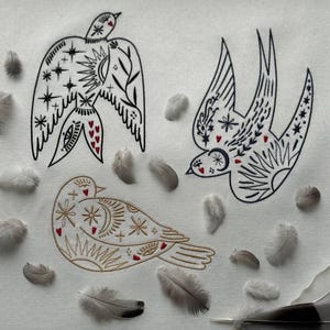Könnte beinhalten: Gestickte Vogeldesigns in Schwarz und Gold mit roten Herzakzenten auf weißem Hintergrund. Die Vögel sind von verstreuten grauen Federn umgeben. Die Designs weisen himmlische und florale Motive auf.