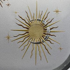 Pode incluir: Desenho bordado de um sol dourado com raios prateados e uma lua crescente, rodeado de pequenas estrelas douradas sobre um fundo de tecido branco.