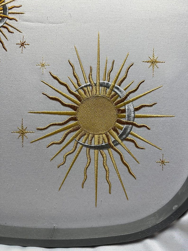 K&ouml;nnte beinhalten: Gestickte Darstellung einer goldenen Sonne mit silbernen Strahlen und einem zunehmenden Mond, umgeben von kleinen goldenen Sternen auf einem wei&szlig;en Stoffhintergrund.