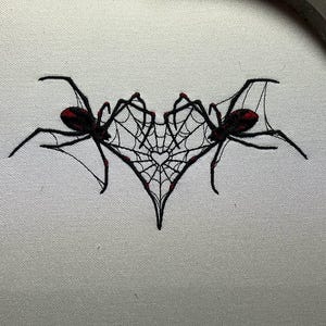 May include: Embroidered design featuring two black spiders with red markings, flanking a heart-shaped web. The spiders and web are stitched on a light-colored fabric, creating a striking contrast. The design is likely for a Halloween or gothic-themed project.