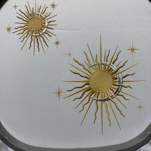 K&ouml;nnte beinhalten: Gestickte Designs mit zwei Sonnen mit Sichelmonden und Sternen auf einem wei&szlig;en Stoffhintergrund. Die Sonnen sind golden mit silbernen Sichelmonden. Die Sterne sind golden.