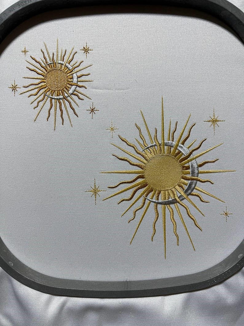 K&ouml;nnte beinhalten: Gestickte Designs mit zwei Sonnen mit Sichelmonden und Sternen auf einem wei&szlig;en Stoffhintergrund. Die Sonnen sind golden mit silbernen Sichelmonden. Die Sterne sind golden.
