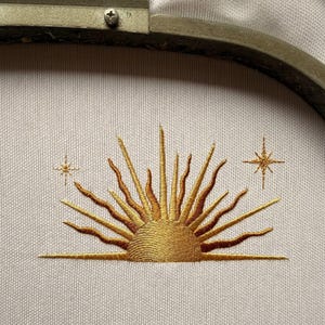 May include: Embroidered design of a golden sun with radiating rays and two starbursts on a light-colored fabric. The embroidery is held in a metal hoop, perfect for a craft project or decorative piece.