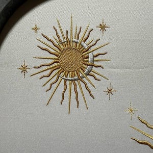 K&ouml;nnte beinhalten: Gestickte Darstellung einer Sonne und eines Mondes mit einer Sichelmondform und strahlenden Strahlen. Die Sonne und der Mond sind in goldfarbenem Garn gestickt, der Sichelmond in silberfarbenem Garn. Um Sonne und Mond sind kleine Sterne in goldfarbenem Garn verteilt.