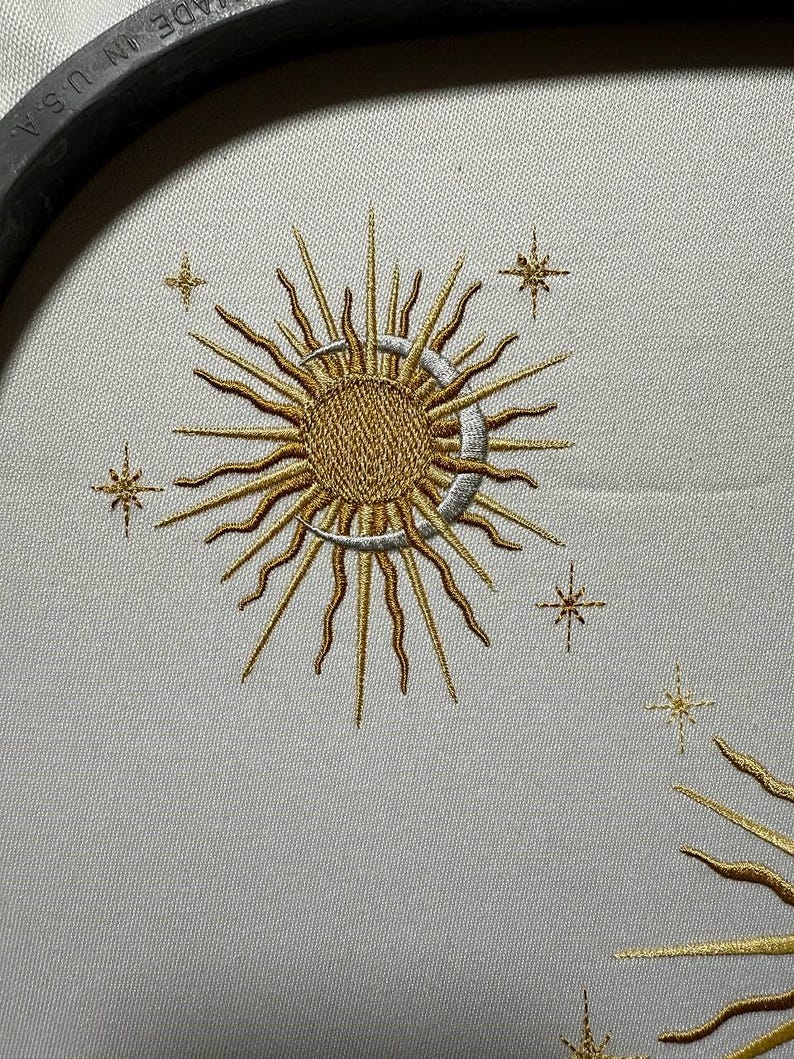 K&ouml;nnte beinhalten: Gestickte Darstellung einer Sonne und eines Mondes mit einer Sichelmondform und strahlenden Strahlen. Die Sonne und der Mond sind in goldfarbenem Garn gestickt, der Sichelmond in silberfarbenem Garn. Um Sonne und Mond sind kleine Sterne in goldfarbenem Garn verteilt.