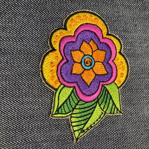 May include: A colorful embroidered flower patch with yellow, orange, pink, purple, and green thread. The flower has a five-petal design with a smaller circle in the center.