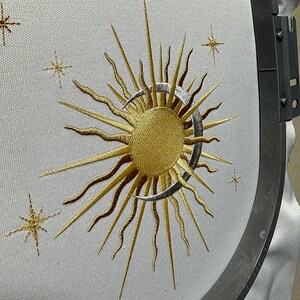 K&ouml;nnte beinhalten: Gestickte Darstellung einer Sonne und eines Mondes mit einer Sichelmondform. Die Sonne ist golden und der Mond ist silbern. Das Design ist von kleinen Sternen umgeben.