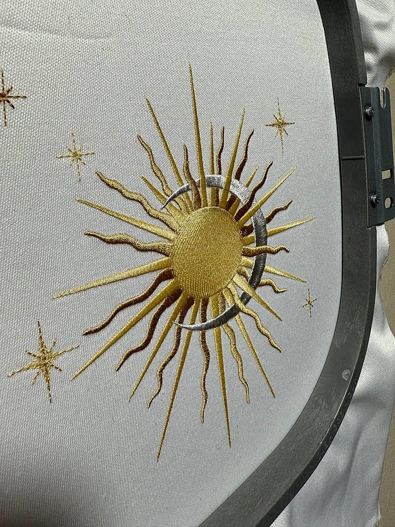 K&ouml;nnte beinhalten: Gestickte Darstellung einer Sonne und eines Mondes mit einer Sichelmondform. Die Sonne ist golden und der Mond ist silbern. Das Design ist von kleinen Sternen umgeben.