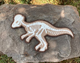 May include: A brown fabric patch with a white embroidered dinosaur skeleton. The dinosaur is in a running pose with its head turned to the side.