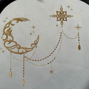 Puede incluir: Diseño bordado con una luna creciente dorada con detalles intrincados, conectada por cadenas de cuentas a una estrella y colgantes en forma de lágrima. Estrellas adicionales están dispersas alrededor de la luna y la estrella. El diseño está sobre una tela gris claro.