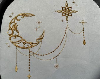 Könnte beinhalten: Gesticktes Design mit einem goldenen Halbmond mit komplizierten Details, der durch Perlenketten mit einem Stern und tropfenförmigen Anhängern verbunden ist. Zusätzliche Sterne sind um Mond und Stern verstreut. Das Design befindet sich auf einem hellgrauen Stoff.