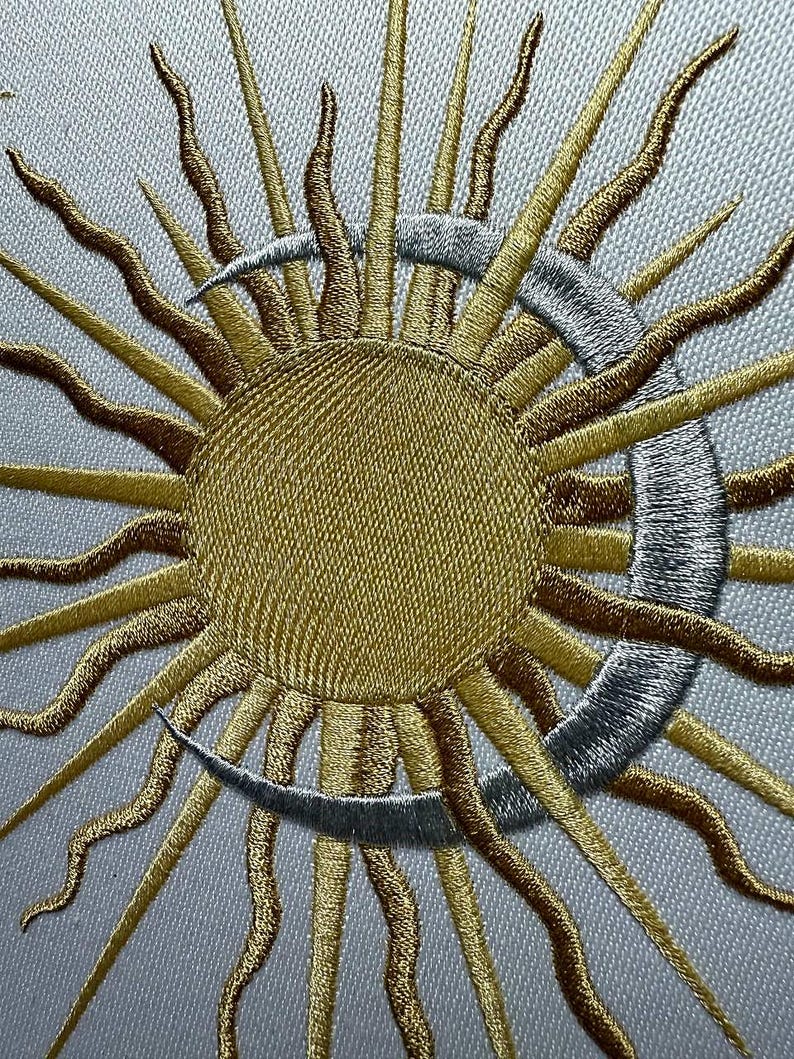K&ouml;nnte beinhalten: Gestickte Sonnen- und Mond-Darstellung mit gold- und silberfarbenem Garn. Die Sonne hat eine gro&szlig;e, runde Mitte und viele Strahlen. Der Mond ist halbmondf&ouml;rmig.