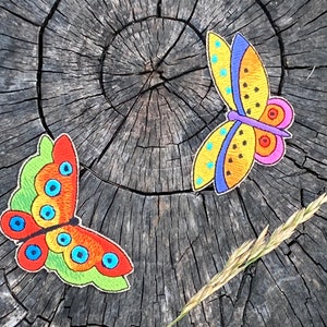 May include: Two embroidered butterfly patches on a wooden surface. One butterfly is yellow with black and blue accents, the other is red, orange, and green with blue eyes.