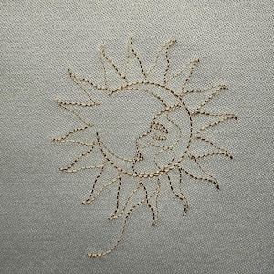 Op de afbeelding: Een gouden geborduurd ontwerp van een zon en maan op een lichtgrijze stof.