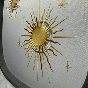 K&ouml;nnte beinhalten: Gestickte Darstellung einer goldenen Sonne und eines Mondes mit einer Sichelform, umgeben von kleineren goldenen Sternen auf einem wei&szlig;en Stoff.