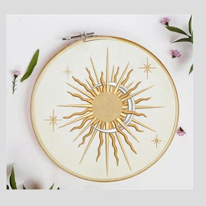 K&ouml;nnte beinhalten: Ein wei&szlig;er Stickrahmen mit einem goldenen Sonnen- und Mondmotiv. Die Sonne hat eine strukturierte Mitte und ist von Strahlen umgeben. Der Mond ist halbmondf&ouml;rmig. Um Sonne und Mond befinden sich vier kleine goldene Sterne.