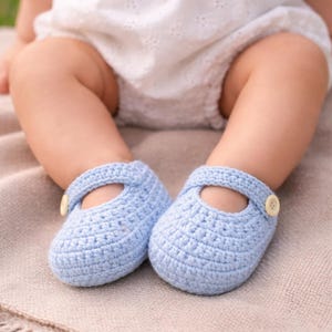 Puede incluir: Un par de zapatos Mary Jane de ganchillo azul bebé con cierre de botón. Los zapatos están hechos a mano y descansan sobre una manta de color neutro. Los zapatos son de color azul claro.