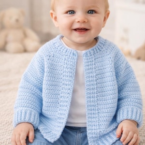 May include: A baby wearing a pale blue, open-front crochet cardigan and denim shorts. The cardigan has a textured stitch pattern and ribbed cuffs. The baby is smiling, with a soft background.