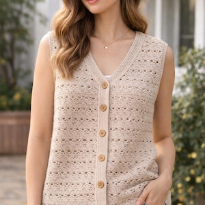 May include: A sleeveless, beige crochet vest with a V-neck and wooden buttons. The vest features a delicate, openwork design and is worn over a white top. The model is wearing blue jeans and a gold necklace.