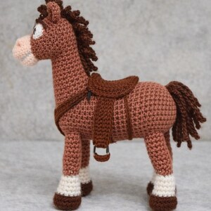 Op de afbeelding: Een gehaakt bruin paard met een zadel, manen en staart. Het paard heeft witte accenten op de hoeven en een lichtroze snuit. Het zadel en het harnas zijn een donkerdere bruintint. Het paard staat rechtop.
