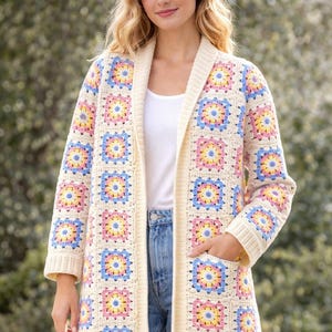 Op de afbeelding: Crèmekleurig gehaakt vest met een kleurrijk bloemmotief. De trui met lange mouwen heeft een sjaalkraag en twee voorzakken. Het bloemmotief bestaat uit roze, gele en blauwe vierkanten. Het vest wordt gedragen over een witte top en een blauwe jeans.