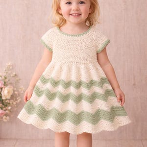 Peut inclure: Robe en crochet crème avec un motif chevron vert sur la jupe. La robe a des manches courtes et une bordure verte autour de l'encolure et des manches. Un nœud vert est dans les cheveux de l'enfant.