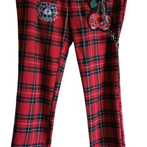 May include: Red tartan trousers with black and green detailing. The trousers have a skull and floral patch, a cherry charm, and black lace trim. A distinctive and fashionable garment.