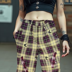 Op de afbeelding: Geel en paars geruite broek met vogel- en decoratieve details. De broek heeft zakken en een ketting. Het model draagt een zwarte crop top en heeft tatoeages op de armen.