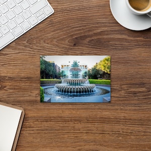 May include: A photograph of a tiered fountain with water cascading down into a circular pool. The fountain is surrounded by greenery and buildings. The image is on a wooden surface with a keyboard and coffee cup.