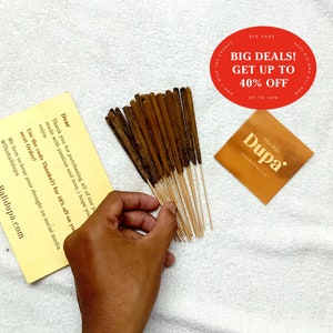 Op de afbeelding: Een bundel bruine wierookstokjes met houten houders. De wierookstokjes zijn in een waaier vorm gerangschikt en worden in een hand gehouden. De achtergrond is een witte stof. De tekst "The Bali Dupa" is zichtbaar op een bruin papieren etiket.