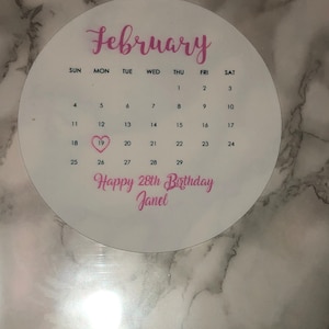 May include: A white calendar with the month of February printed on it. The 19th is highlighted with a pink heart and the text "Happy 28th Birthday Janel" is printed below the calendar.