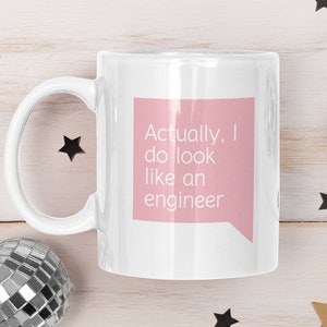 May include: White ceramic mug with a pink speech bubble that says "Actually, I do look like an engineer". The mug is on a white wooden surface with gold and black confetti.