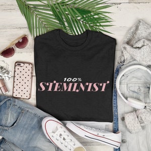 Könnte beinhalten: Ein schwarzes T-Shirt mit dem Text "100% STEMINIST" in rosa Schrift. Das Shirt liegt auf einer weißen Holzoberfläche mit einer Jeans, einer Jeansjacke, einer Sonnenbrille, einer Handyhülle und einem Kopfhörer.