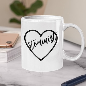 May include: White ceramic mug with a black heart design and the word "Steminist" in a handwritten font. The mug has a curved handle and is sitting on a marble surface. A smartphone, a stack of white paper, and a pen are also visible.