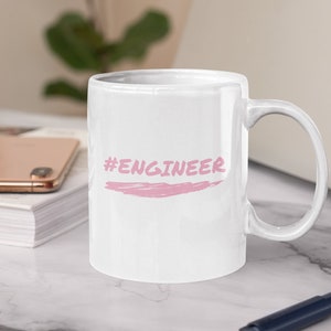 Könnte beinhalten: Weiße Keramiktasse mit einem rosafarbenen Hashtag und dem Wort "ENGINEER" in rosafarbener Pinselschrift.