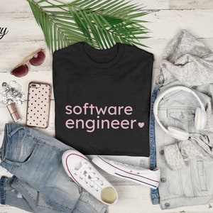 Könnte beinhalten: Ein schwarzes T-Shirt mit dem Text "software engineer" in rosa Schrift und einem rosa Herz. Das Shirt liegt auf einem weißen Holzgrund mit einer Jeans, einer Jeansjacke, einer Sonnenbrille, einem Telefon und Kopfhörern.