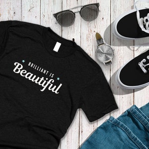 Könnte beinhalten: Ein schwarzes T-Shirt mit dem Text "BRILLIANT IS BEAUTIFUL" in weißen Buchstaben. Das Shirt liegt auf einer weißen Holzoberfläche mit einem Paar schwarzer Sneakers, einer Sonnenbrille und einer Uhr.