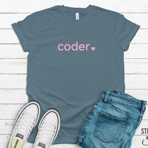 Può includere: Una maglietta blu con la parola "coder" in lettere rosa e un cuore rosa. La maglietta è appoggiata su una superficie di legno bianco con un paio di scarpe da ginnastica bianche e un paio di jeans blu.