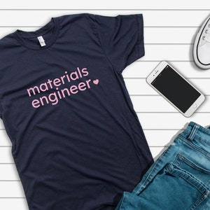 May include: A navy blue t-shirt with the text "materials engineer" in pink lettering and a pink heart. The shirt is laid out on a white wood surface with a pair of white trainers, blue jeans, and a smartphone.