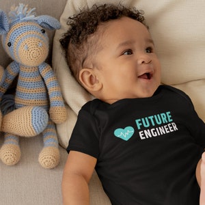 以下が含まれることがあります： 「FUTURE ENGINEER」という文字と、心臓の鼓動のグラフィックが描かれたターコイズブルーのハートがプリントされた黒いボディスーツを着た赤ちゃん。赤ちゃんは、2つの編みぐるみのおもちゃと一緒に、薄い茶色のソファに寝転んでいます。