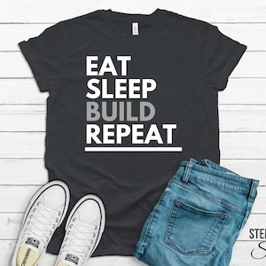 Peut inclure: Un t-shirt gris foncé avec le texte "Eat Sleep Build Repeat" en lettres blanches et grises. Le t-shirt est présenté sur un fond en bois blanc avec une paire de baskets blanches et un jean bleu.